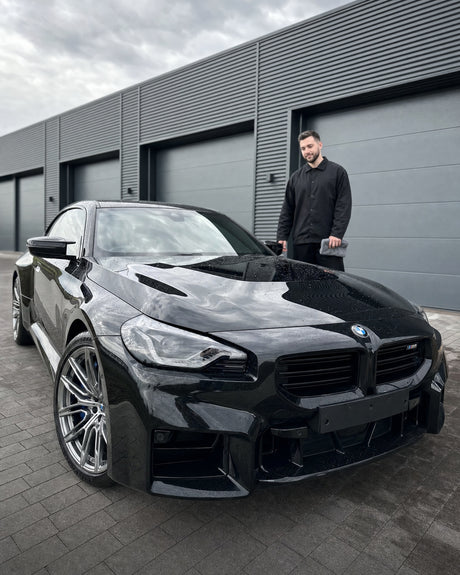 Daniel mit Mikrofasertuch neben dem glänzend schwarzen BMW M2 (G87), die Frontscheibe spiegelt streifenfrei klar.