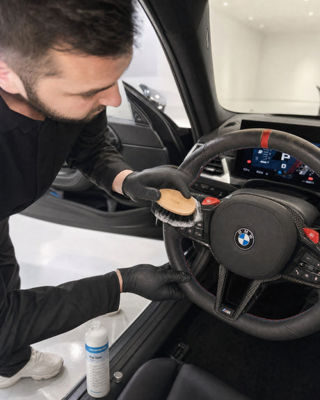 Daniel von Detailing1 arbeitet Reinigungsschaum mit einer weichen Lederbürste in den Lenkradkranz eines weißen BMW M2 ein — g